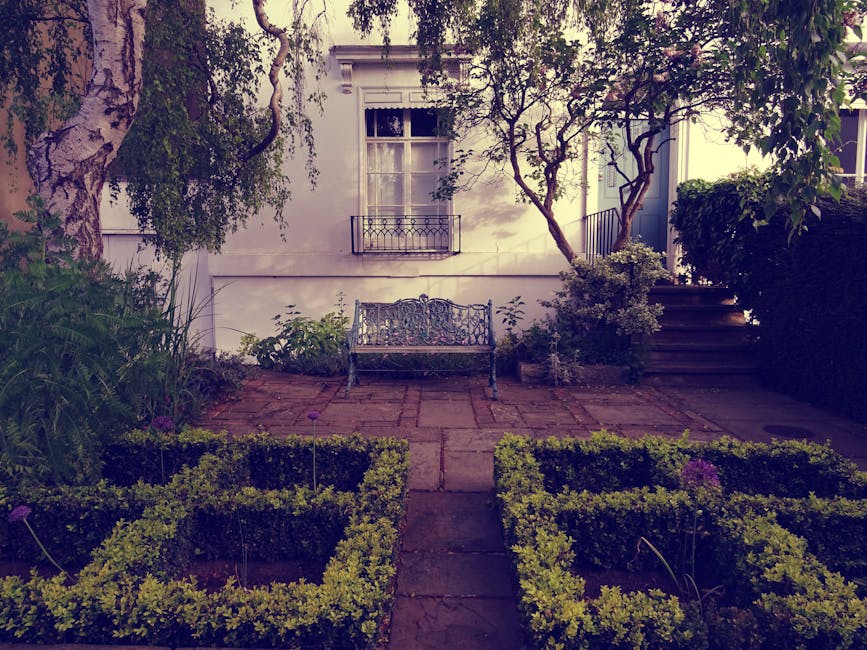 A small, neatly maintained garden scene featuring a paved patio area with a decorative metal bench positioned against a white exterior wall of a house. The garden is bordered by low, square-shaped box