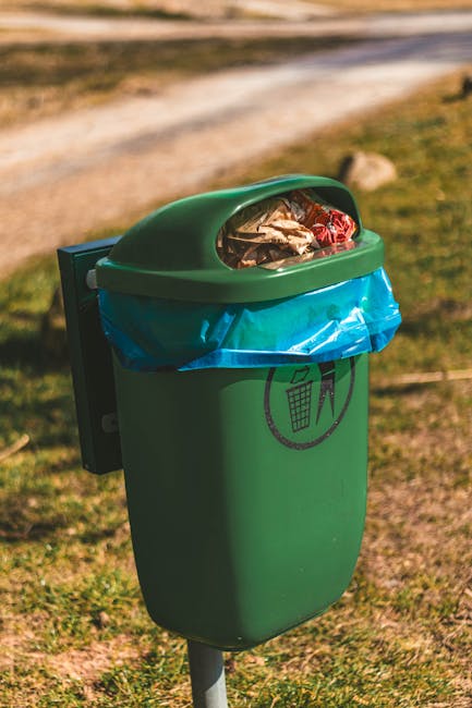 A person wearing a light-colored glove and a watch is collecting garden waste, including small branches, leaves, and twigs, into a white open-ended sack using a shovel. The garden waste consists of tr